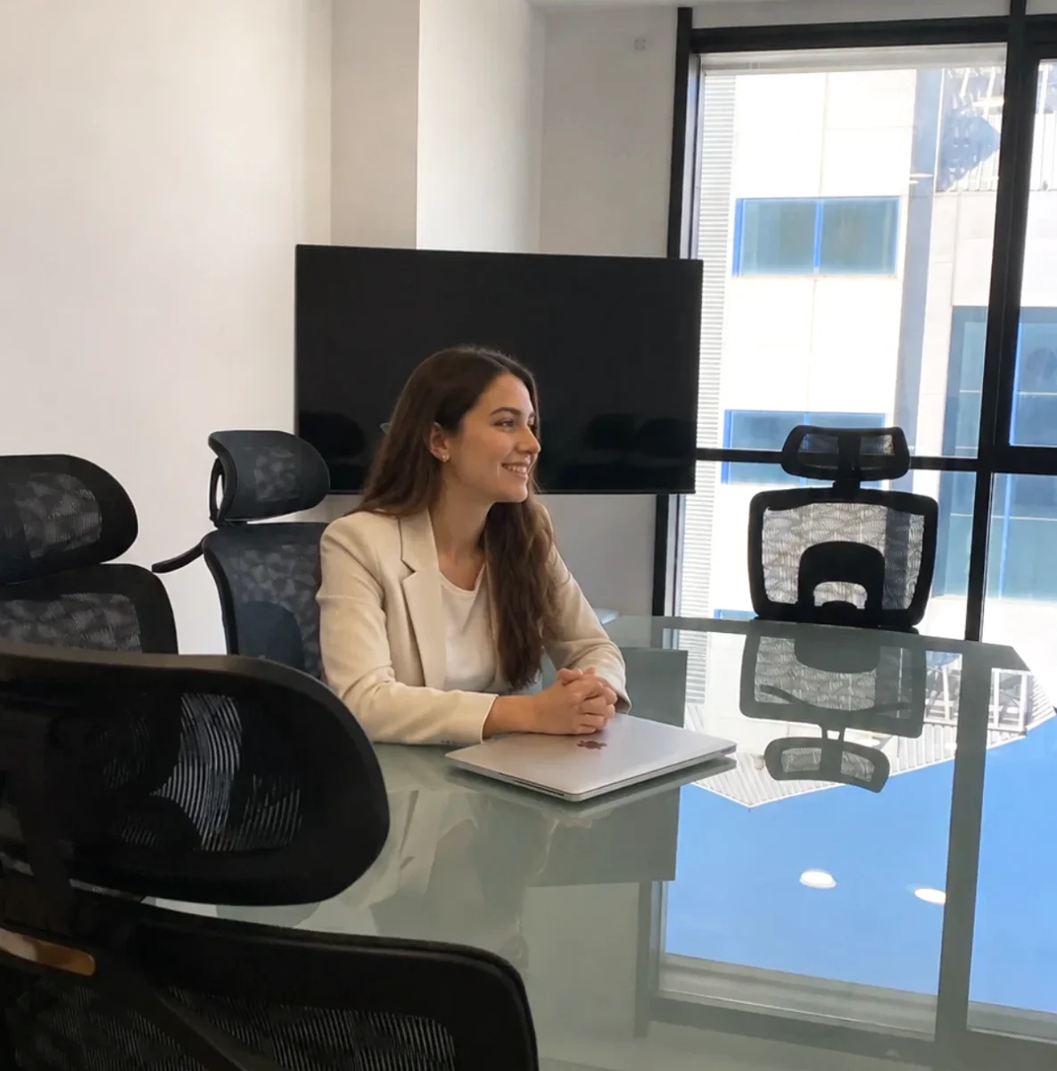 Profesional sonriendo en sala de reuniones con mesa de cristal, sillas ergonómicas, pantalla y ventanal con luz natural